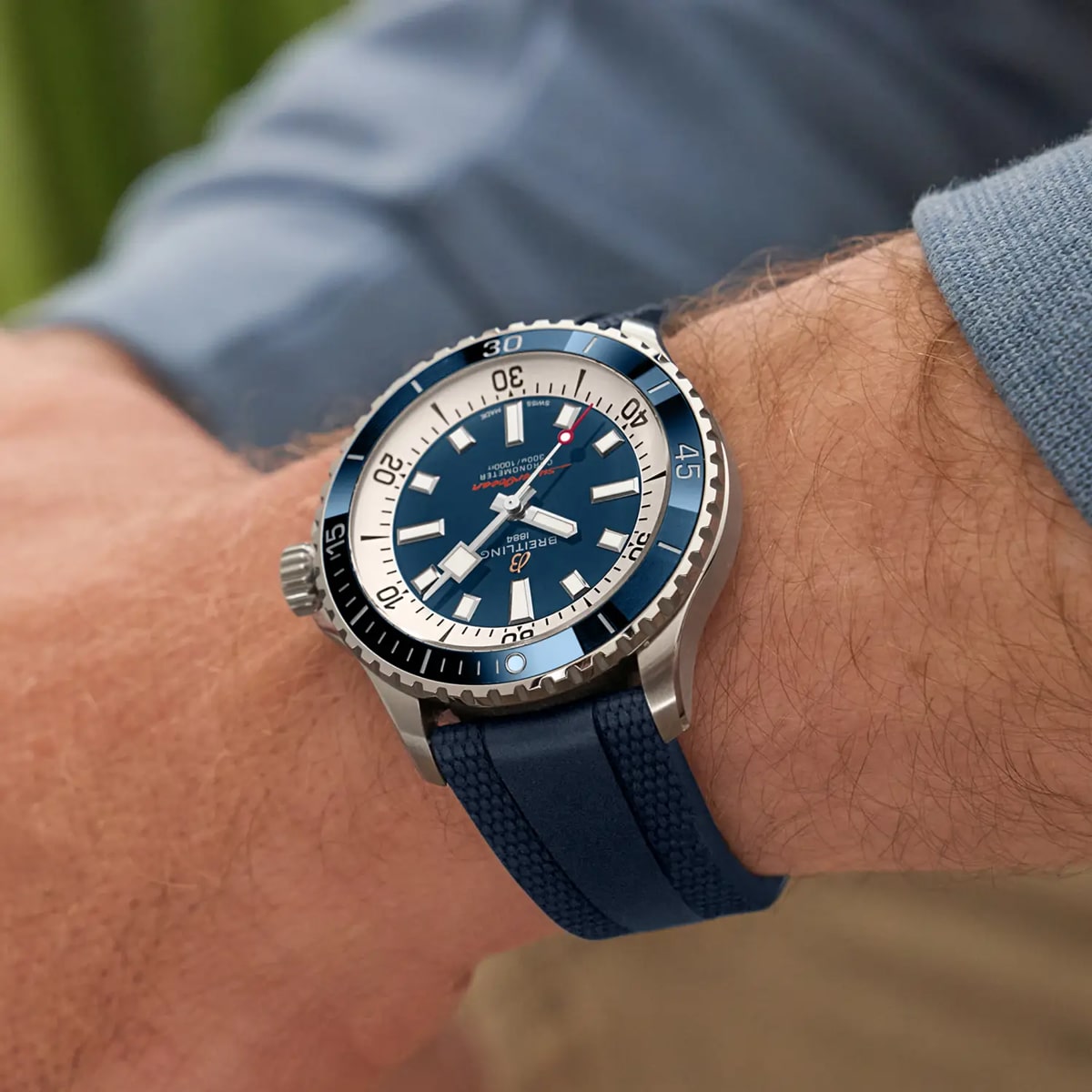 Eine Nahaufnahme des Handgelenks einer Person, die eine blaue Taucheruhr aus der Breitling Superocean Kollektion mit blauem Armband, silberner Lünette und weißen Stundenmarkierungen trägt. Die Person trägt ein blaues langärmeliges Hemd vor einem unscharfen Hintergrund.