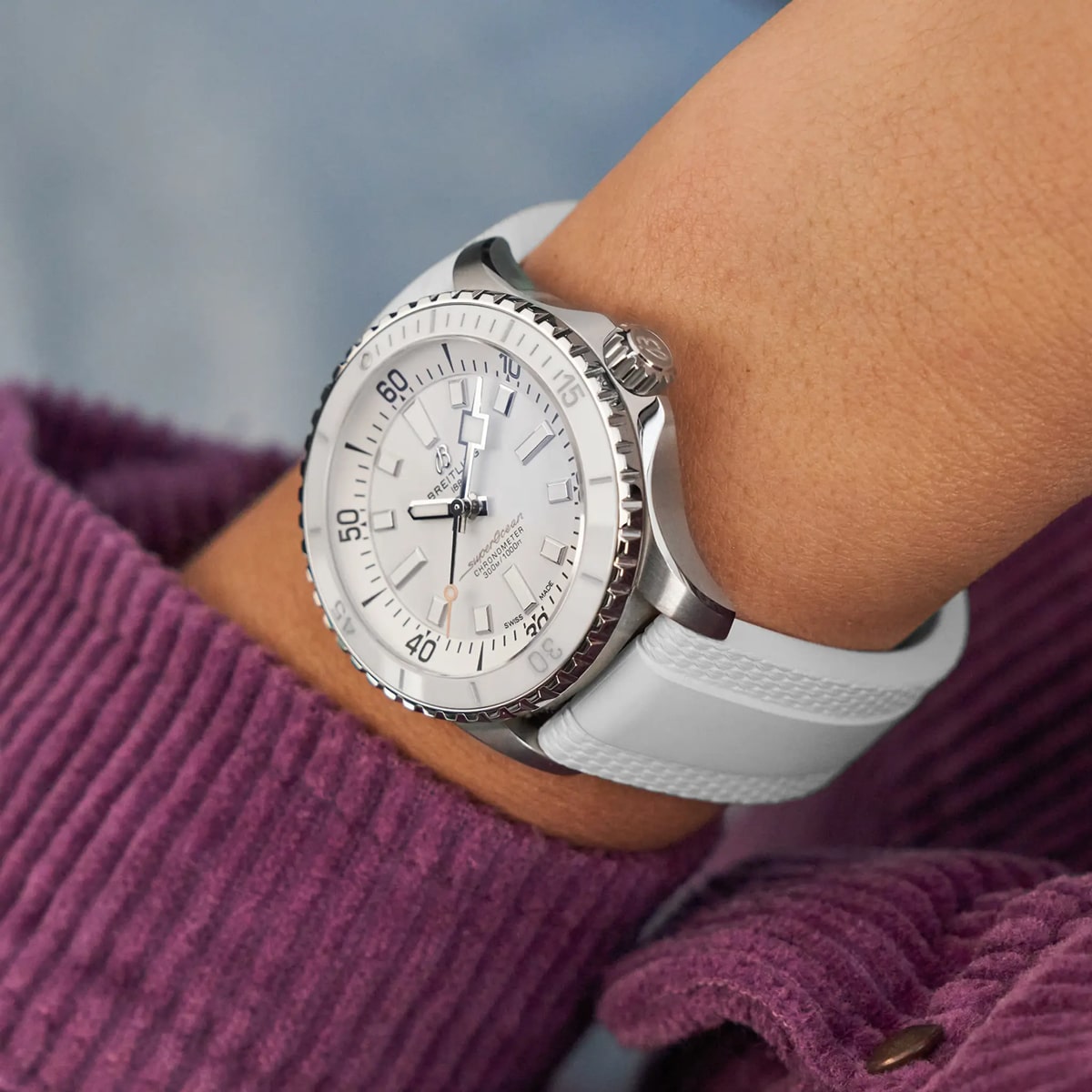 Eine Nahaufnahme des Handgelenks einer Person, die eine silber-weiße Taucheruhr der Breitling Superocean Kollektion mit weißem Kautschukarmband trägt. Die Person ist mit einer lila Cordjacke und einem hellbraunen Oberteil bekleidet.