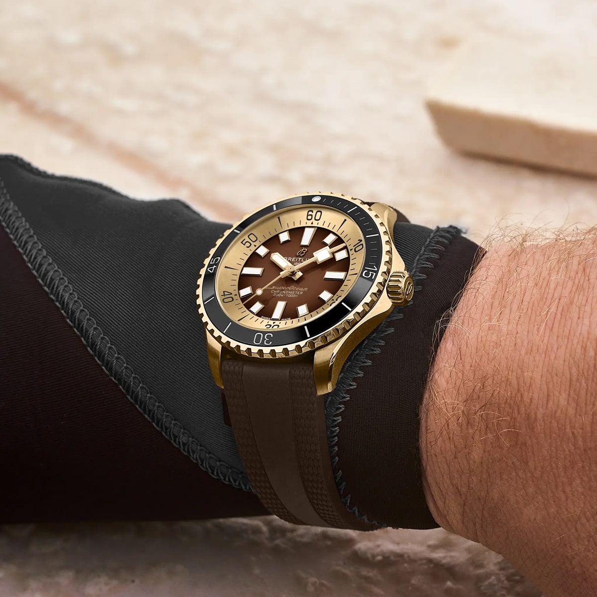 Eine goldene und braune Armbanduhr der Breitling Superocean Kollektion mit drehbarer Lünette und Kautschukarmband wird am Handgelenk einer Person getragen, die in eine schwarze Neoprenhülle gekleidet ist. Der Hintergrund ist unscharf und zeigt eine helle Oberfläche.