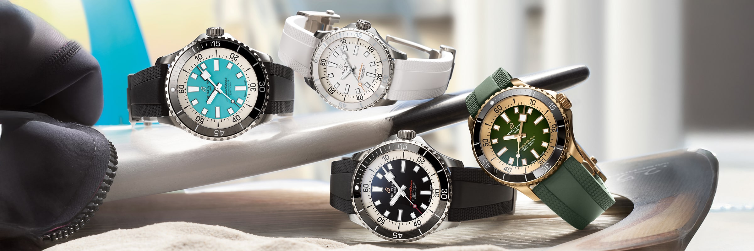 Vier Armbanduhren der Breitling Superocean Kollektion mit türkisem, weißem, schwarzem und grünem Zifferblatt liegen auf einem Tisch mit passenden Armbändern, einem silbernen und goldenen Stift und einem schwarzen Objekt im Hintergrund.