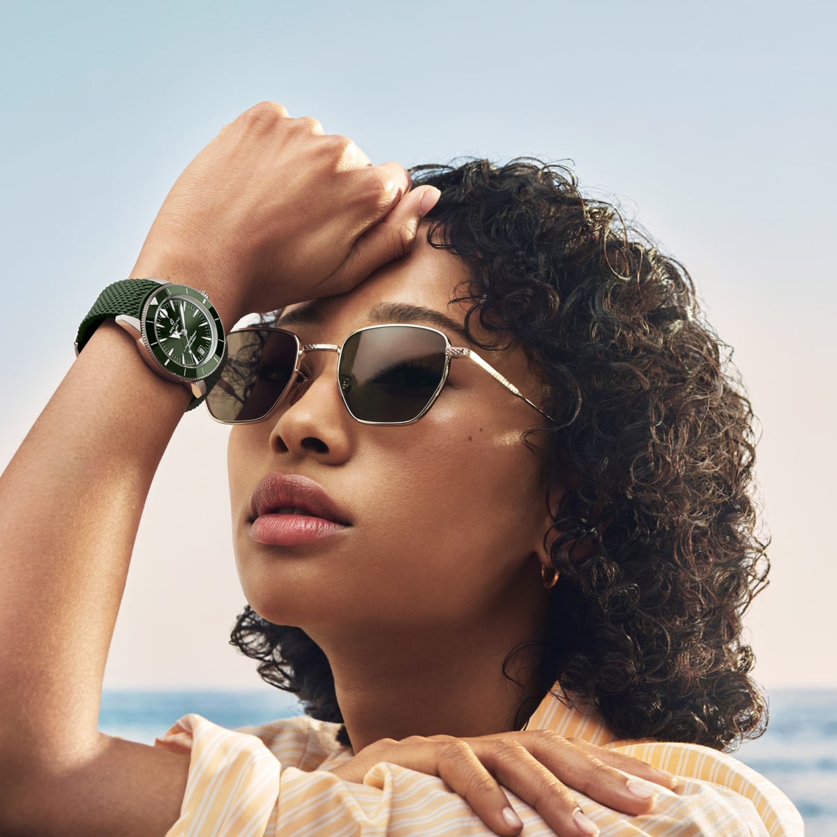 Eine Frau mit Sonnenbrille und einer grünen Breitling Super Ocean Heritage Armbanduhr stützt ihre Hand auf ihre Stirn. Sie hat lockiges Haar und befindet sich im Freien mit einem ruhigen, blauen Himmel und dem Meer im Hintergrund.