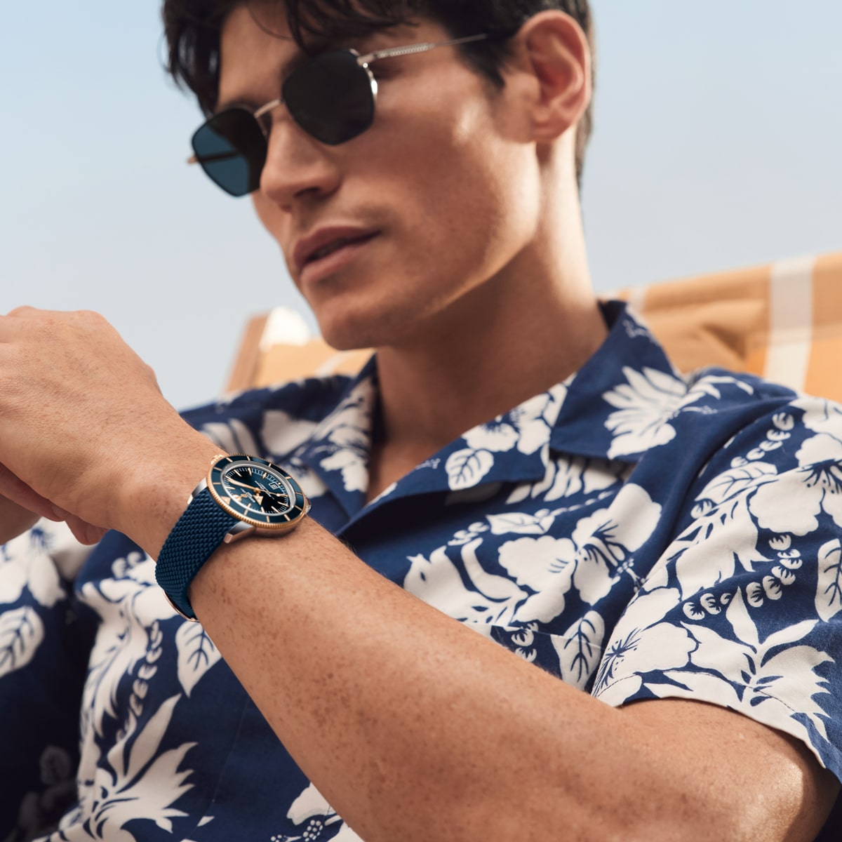 Ein Mann mit Sonnenbrille und blauem geblümten Hemd sitzt im Freien und zeigt eine blaue Breitling Super Ocean Heritage Armbanduhr an seinem linken Handgelenk. Er wirkt entspannt, und der unscharfe Hintergrund deutet auf einen sonnigen Tag hin.