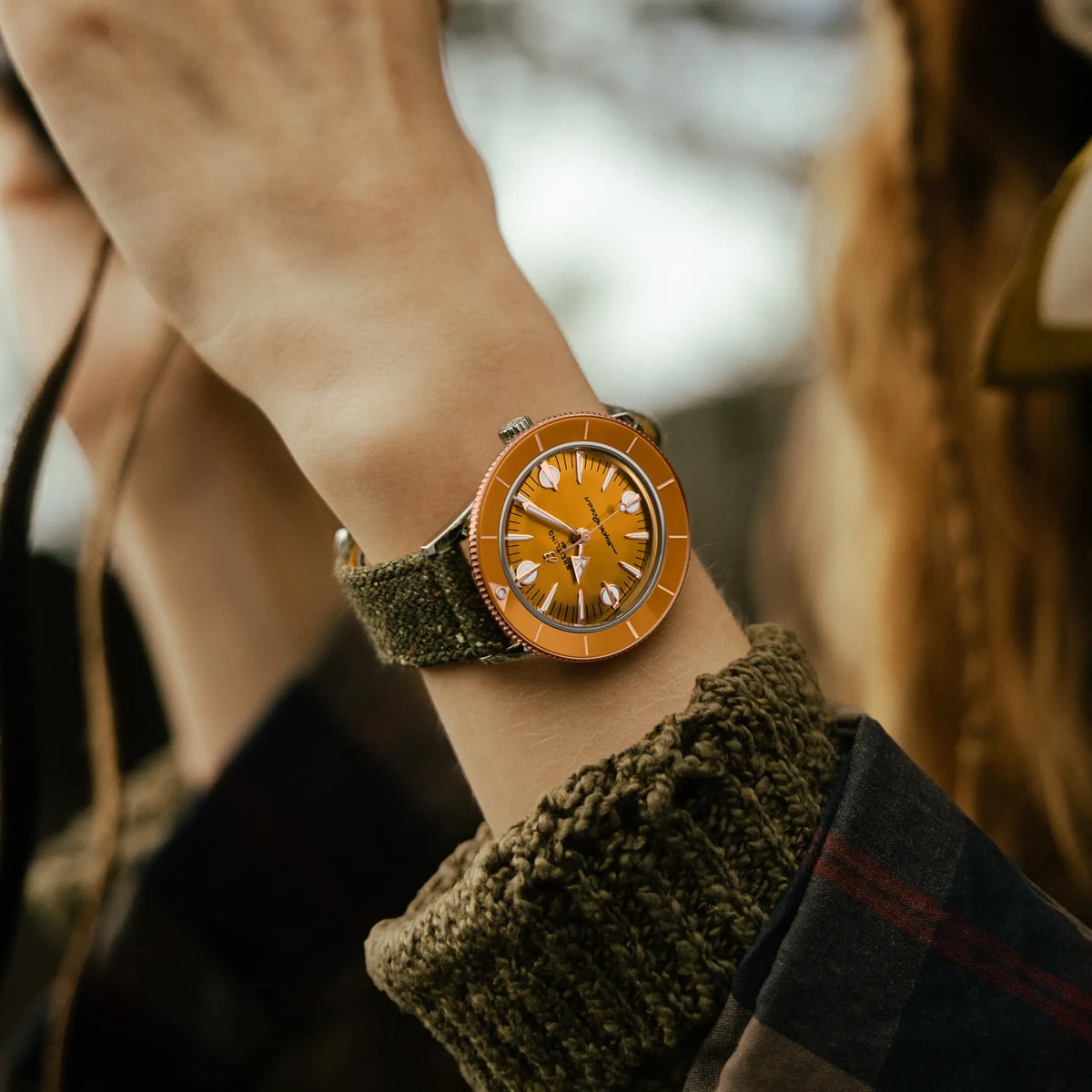 Eine Person, die einen dicken olivgrünen Pullover trägt, hält ihr Handgelenk hoch und zeigt eine orangefarbene Breitling Super Ocean Heritage-Uhr mit einem strukturierten grünen Armband. Der Hintergrund ist leicht unscharf, was auf eine Umgebung im Freien hindeutet.
