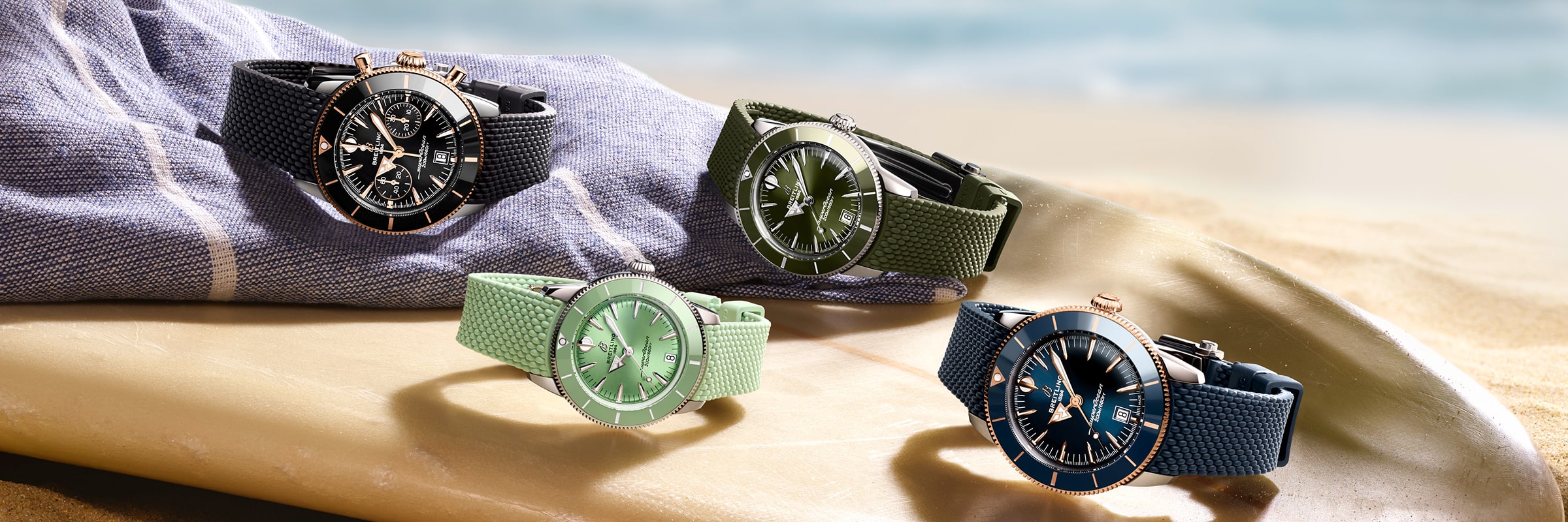 Vier Breitling Super Ocean Heritage Armbanduhren mit schwarzen, olivgrünen, hellgrünen und marineblauen Armbändern liegen an einem Sandstrand auf einem Surfbrett und einem gestreiften Handtuch, während das Meer im Hintergrund verschwimmt.