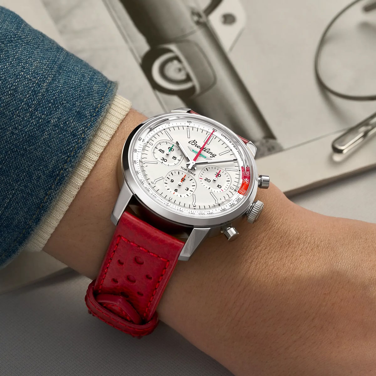 Eine Person trägt eine silberne Chronographenuhr der Breitling Top Time Kollektion mit weißem Zifferblatt und rotem, perforiertem Lederarmband. Der Hintergrund zeigt einen Teil eines Jeansärmels und ein Schwarz-Weiß-Foto eines Oldtimers.
