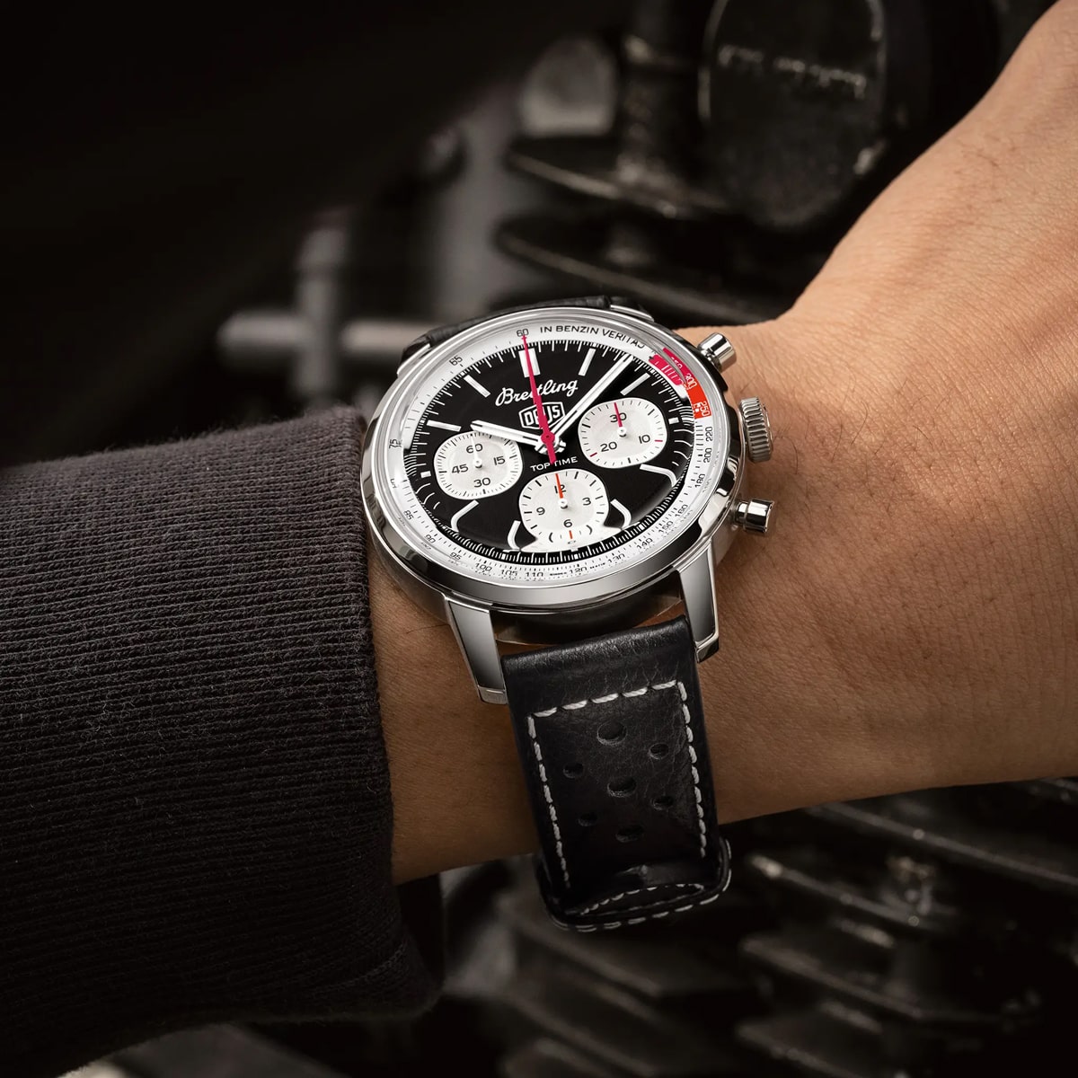 Nahaufnahme eines Handgelenks mit einem Breitling Top Time Kollektion Chronographen mit schwarzem Zifferblatt, drei weißen Hilfszifferblättern und einem schwarzen perforierten Lederarmband mit weißen Nähten. Der unscharfe Hintergrund lenkt den Blick auf die Uhr.