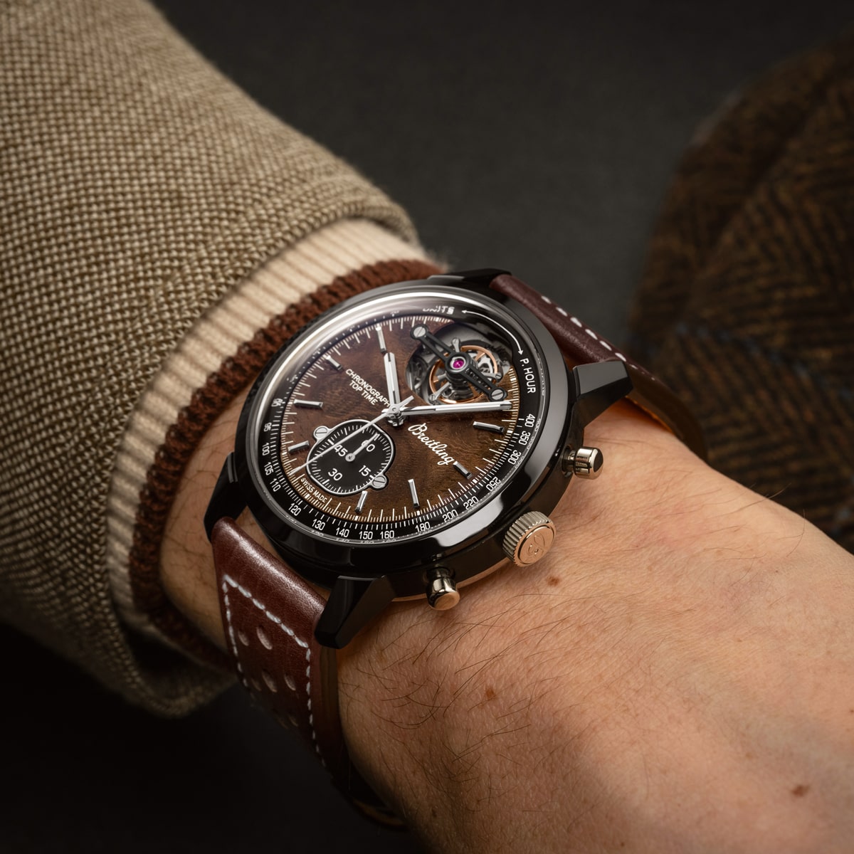 Nahaufnahme einer Person, die eine braune Armbanduhr mit Lederarmband aus der Breitling Top Time Kollektion mit detailliertem braunem Zifferblatt und sichtbarem Tourbillon-Mechanismus trägt, gekleidet in eine braune Tweedjacke und ein beiges Hemd.