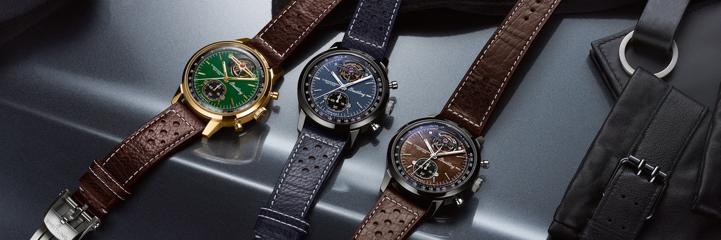 Drei Luxusarmbanduhren der Breitling Top Time Kollektion, jeweils mit Lederarmband, werden nebeneinander auf einer Metalloberfläche präsentiert. Grüne, blaue und braune Zifferblätter bieten ein sportlich-elegantes Design, bei dem ein Lederhandschuh teilweise sichtbar ist.