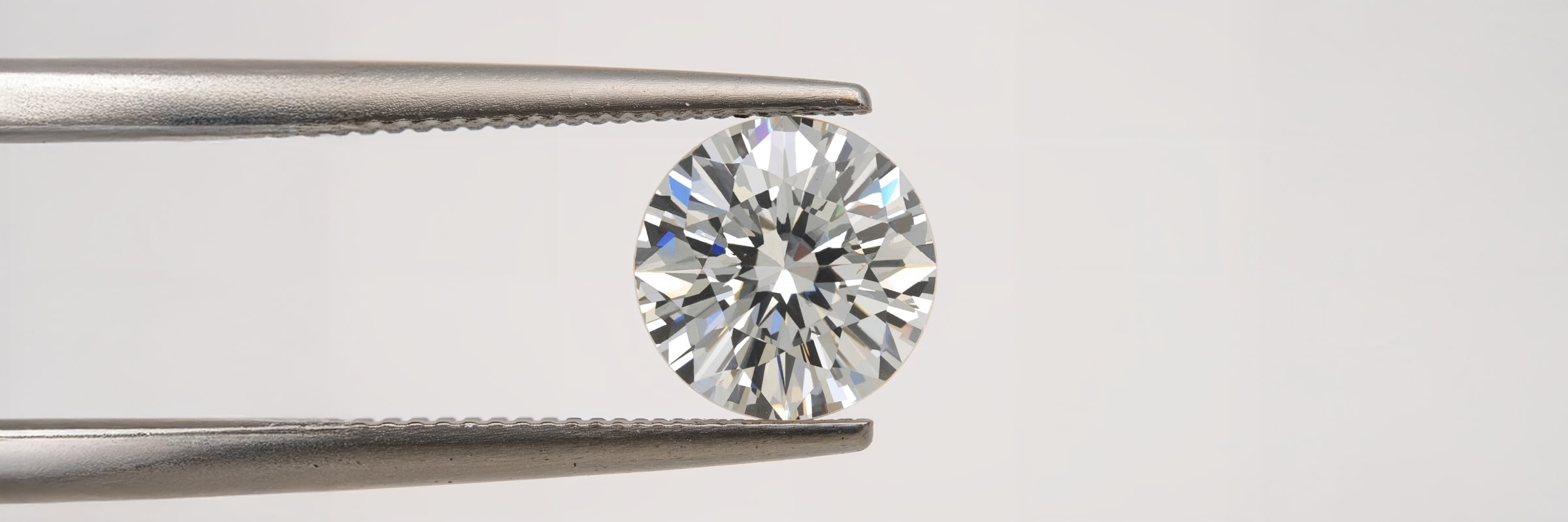 © Juwelier Wagner-Madler e.K. Ein runder, facettierter Brillant wird zart zwischen den Spitzen einer Metallpinzette vor einem schlichten, hellen Hintergrund gehalten.