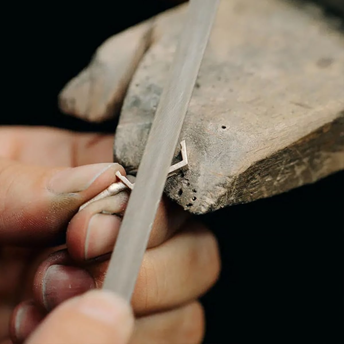 Nahaufnahme der Hand eines Goldschmiedes, der mit einer Metallfeile ein kleines Metallschmuckstück auf einer Holzwerkbank formt. Die Person hält das filigrane Stück vorsichtig und arbeitet mit präzisen Bewegungen an feinen Details. Der Hintergrund ist unscharf.