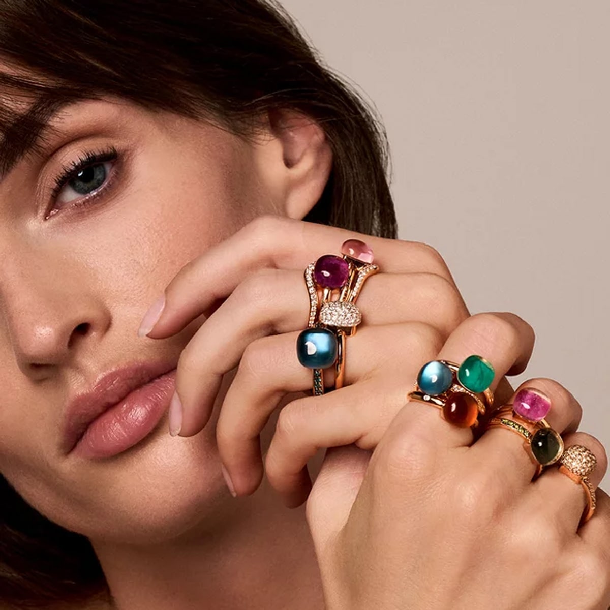 BIGLI © 2026 Eine Frau mit heller Haut und braunem Haar posiert mit ihren Händen in der Nähe ihres Gesichts und präsentiert exquisiten Schmuck - mehrere goldene Ringe, die mit großen, bunten Edelsteinen an ihren Fingern verziert sind.