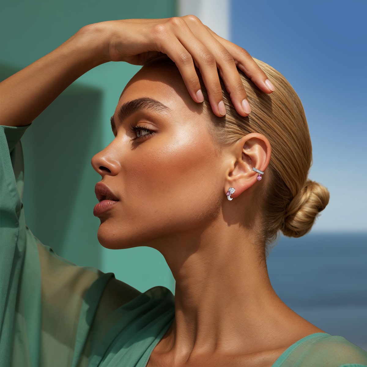 Eine Frau mit blondem Haar in einem tiefen Dutt posiert im Profil, trägt eleganten Silberschmuck und ein durchsichtiges grünes Oberteil. Sie hebt eine Hand an ihre Stirn, umrahmt von einem klaren blauen Himmel und dem Meer im Hintergrund.