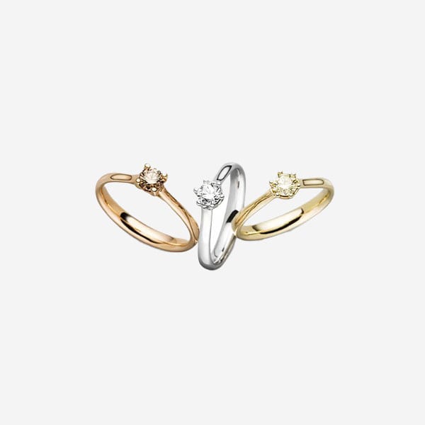 Drei Solitär-Verlobungsringe sind vor einem weißen Hintergrund zu sehen. Die Schmuckringe sind aufrecht in einer Reihe angeordnet; von links nach rechts sind die Ringe aus Roségold, Weißgold und Gelbgold. Jeder Ring ist mit einem Diamanten im Rundschliff in einer klassischen Krappenfassung versehen.