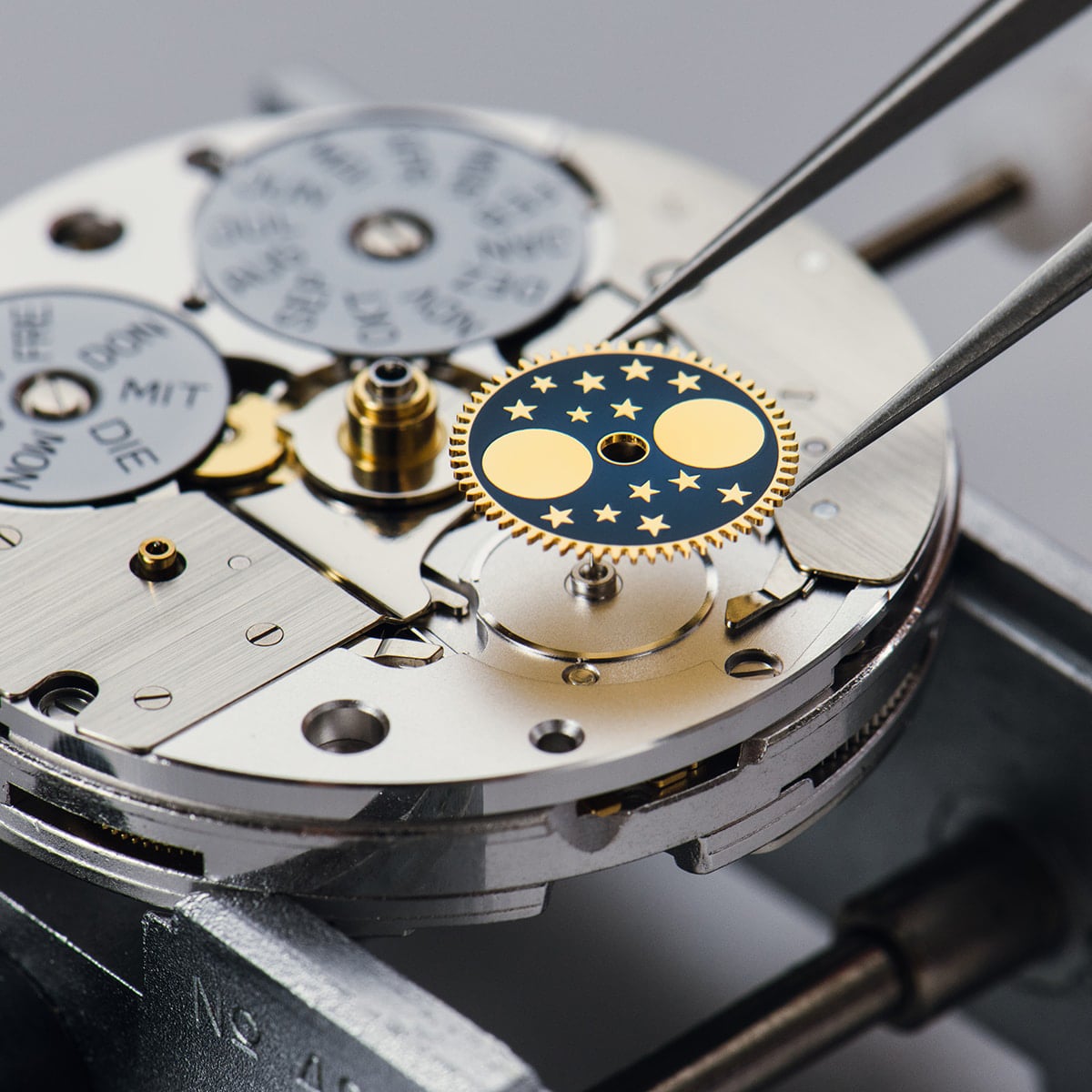 Nahaufnahme des Uhrenmechanismus, während eine Pinzette ein mit Mondphasen und Sternen verziertes Zahnrad vorsichtig auf die komplizierten Metallteile auflegt.