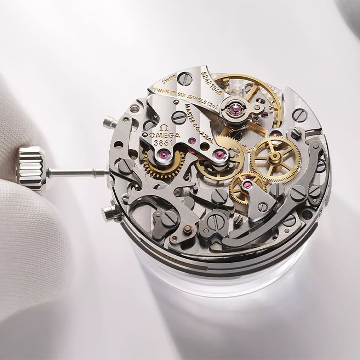 © Juwelier Wagner-Madler e.K. Eine Nahaufnahme der behandschuhten Hand eines Uhrmachers, die ein kompliziertes Omega-Uhrwerk mit sichtbaren Zahnrädern, Edelsteinen und Komponenten in einer fachmännischen Uhrenwerkstatt hält.