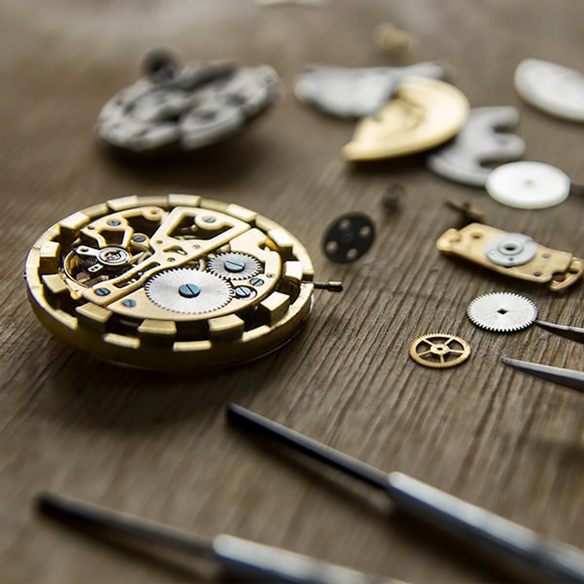 © Juwelier Wagner-Madler e.K. Nahaufnahme von zerlegten Uhrenkomponenten, die auf einer Holzoberfläche ausgebreitet sind. Die komplizierten Zahnräder, Schrauben und Zifferblätter der Uhrenwerkstatt umschließen das Mittelstück und zeigen freiliegende Zahnräder und mechanische Teile. Im Vordergrund sind auch Präzisionswerkzeuge zu sehen.