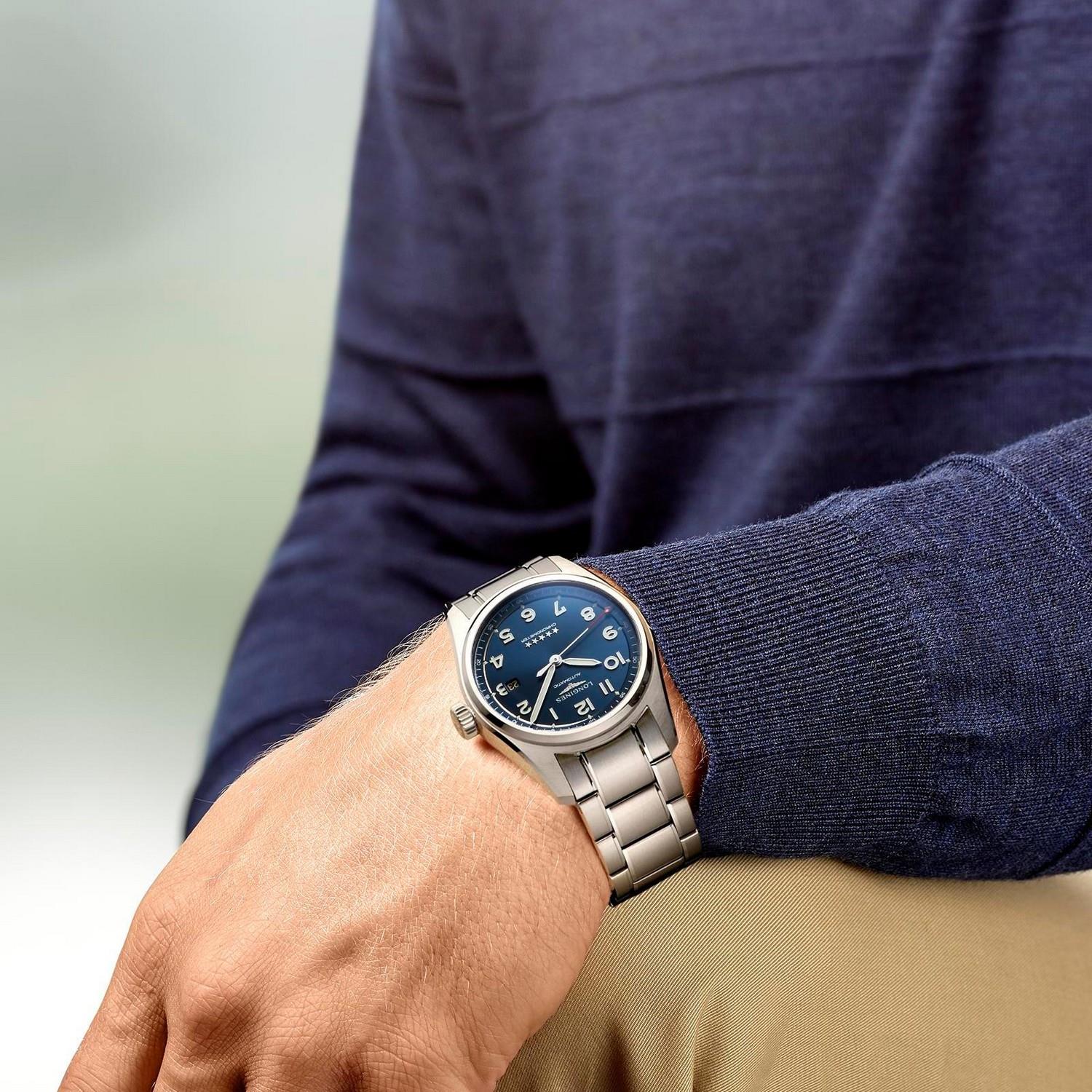 Mit der LONGINES SPIRIT, einer silbernen Armbanduhr mit blauem Zifferblatt und weißen Ziffern, sitzt eine Person in einem marineblauen Pullover und einer beigen Hose und stützt den linken Arm auf das Knie.