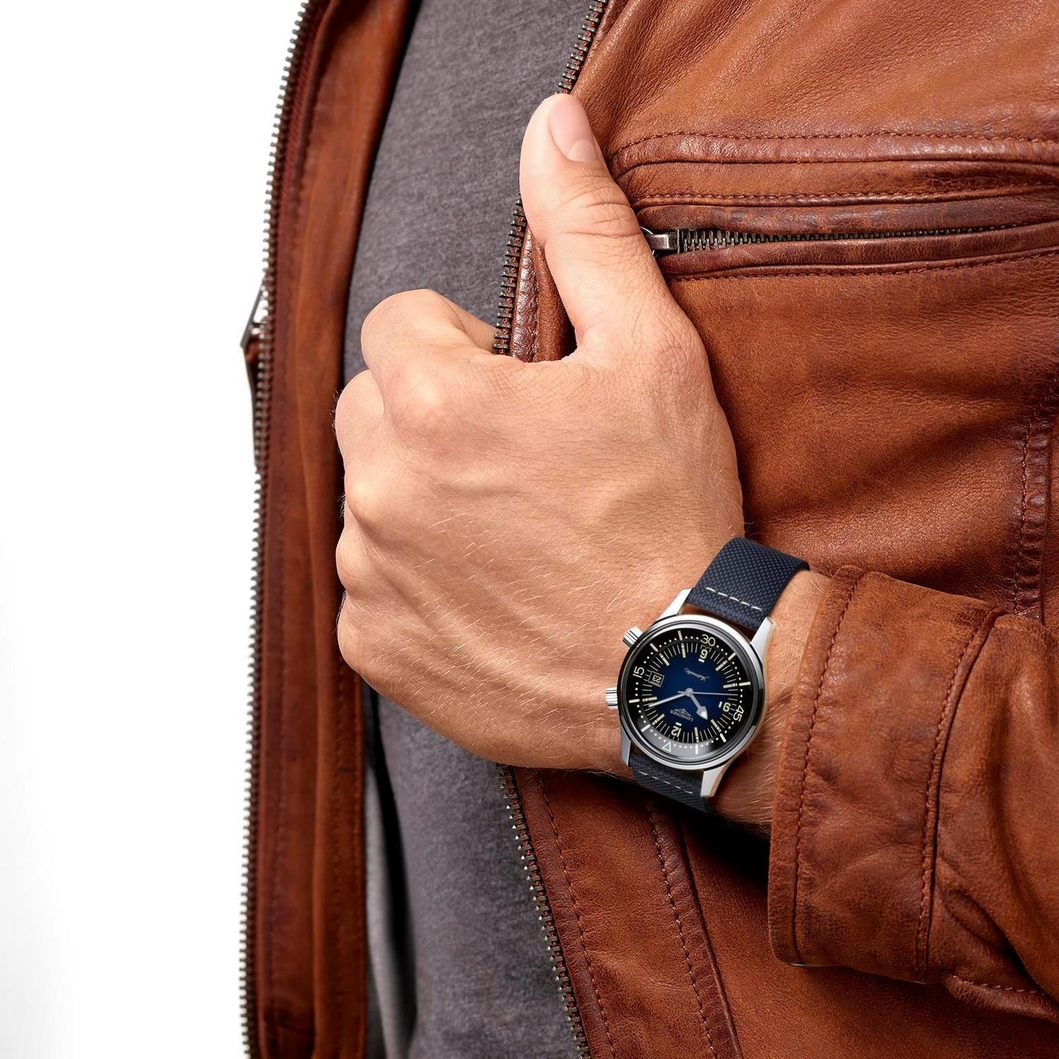 Eine Person in brauner Lederjacke und grauem Hemd zeigt den Daumen nach oben und trägt THE LONGINES LEGEND DIVER WATCH 42MM mit dunkelblauem Zifferblatt und silberner Lünette an ihrem linken Handgelenk.