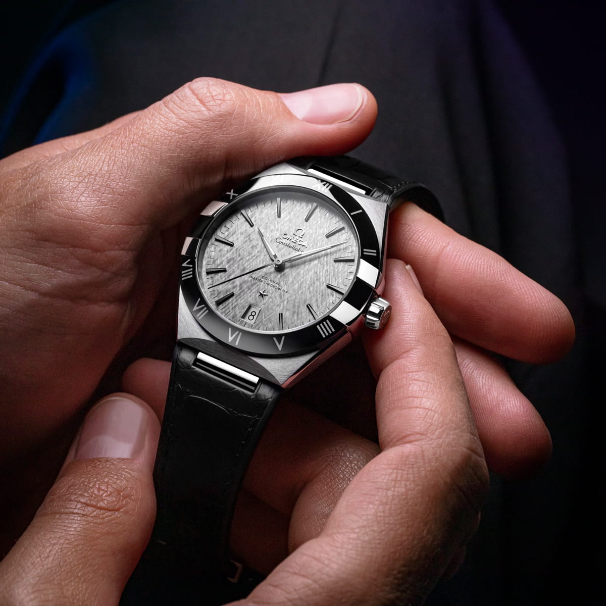 OMEGA © 2026 Eine Nahaufnahme von Händen, die eine OMEGA Constellation Armbanduhr mit silbernem Zifferblatt, schwarzer Lünette und schwarzem Lederarmband halten. Der polierte Zeitmesser zeigt 10:09 vor einem dunklen, verschwommenen Hintergrund.