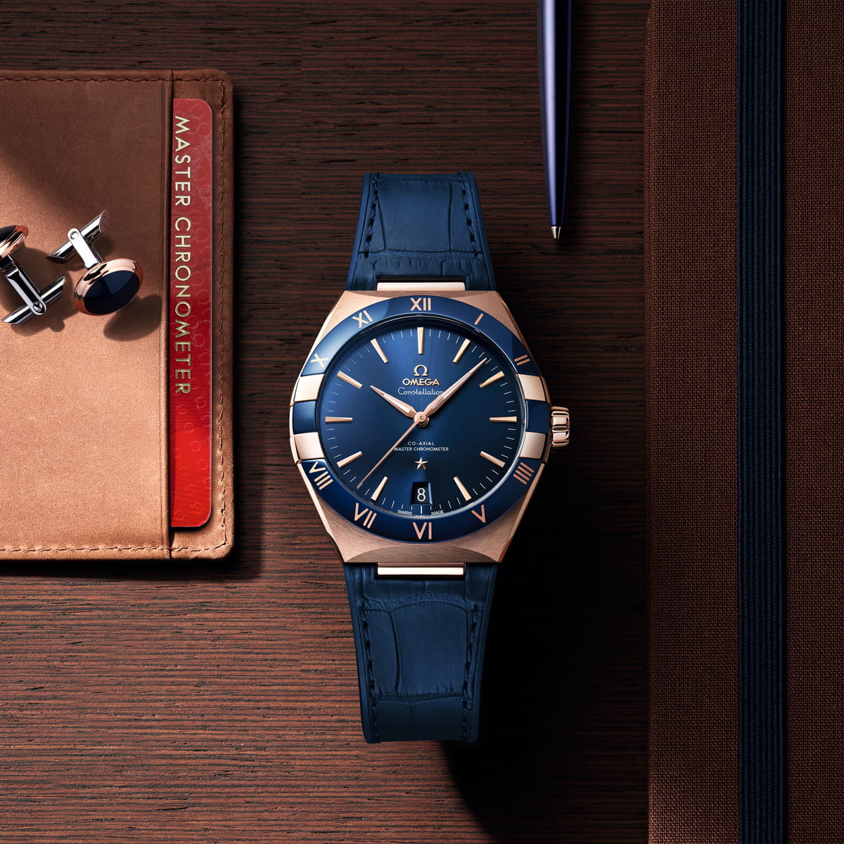 OMEGA © 2026 Eine goldene und blaue OMEGA Constellation Armbanduhr mit blauem Lederarmband liegt auf einem Holztisch neben einem braunen Notizbuch, Manschettenknöpfen, einem Stift und einer roten Master Chronometer-Zertifizierungskarte.