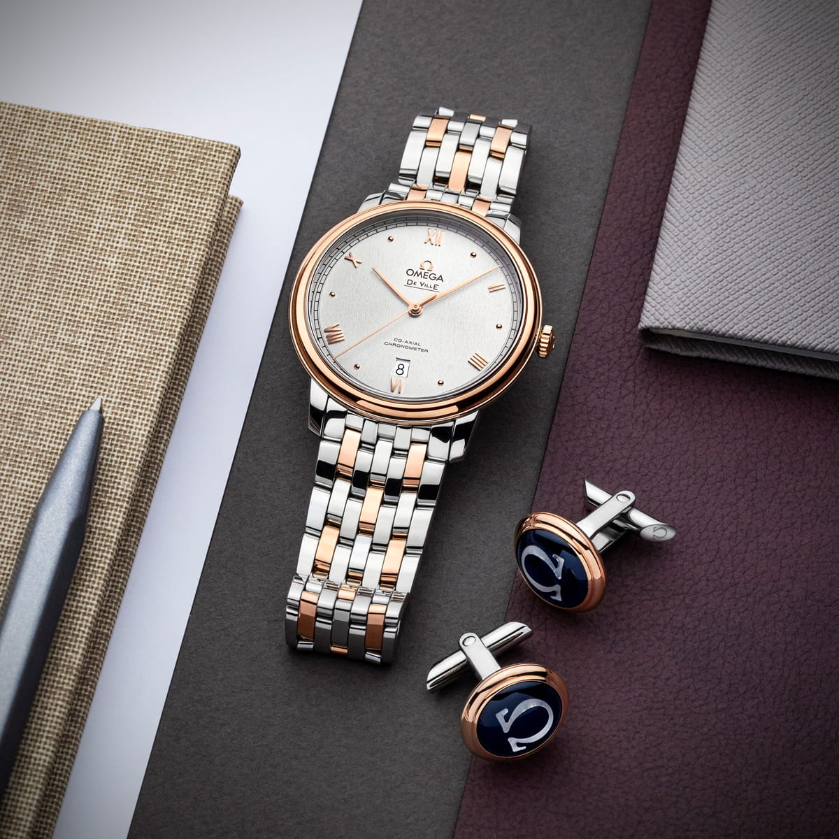 Eine zweifarbige OMEGA DeVille-Armbanduhr mit silberfarbenem und roségoldenem Armband wird auf strukturierten Oberflächen neben einem silbernen Kugelschreiber und marinefarbenen Manschettenknöpfen mit roségoldenen Akzenten präsentiert.