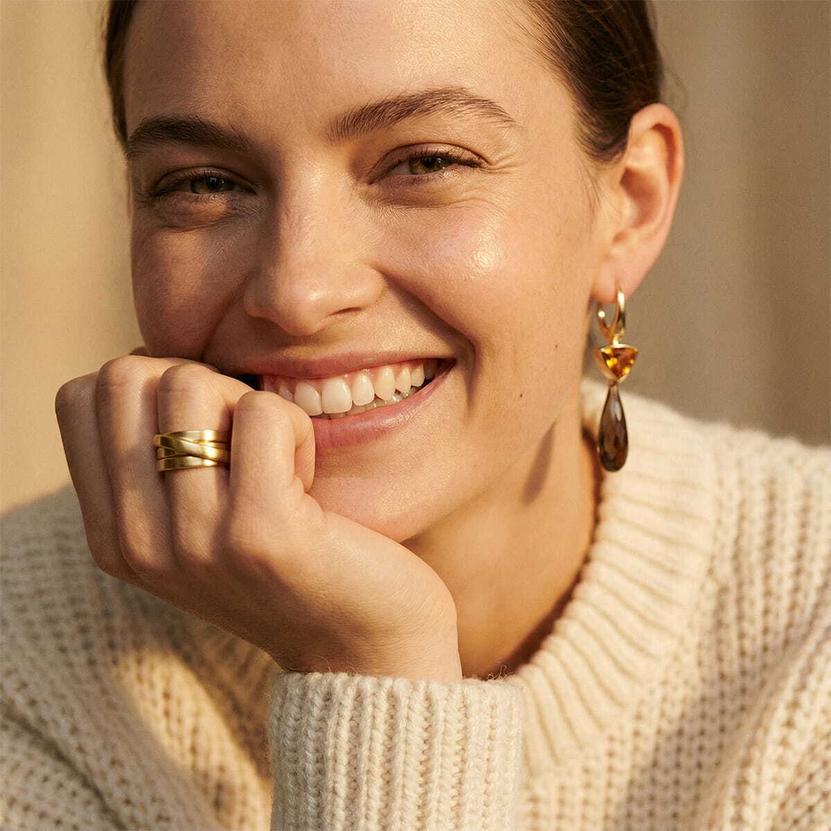 © Juwelier Wagner-Madler e.K. Eine Frau in einem cremefarbenen Strickpullover lächelt warm und zeigt ihre Zähne. Sie stützt ihr Kinn auf ihre Hand, in der sie Goldschmiede-Goldringe und Tropfenohrringe trägt. Die Beleuchtung ist sanft und natürlich und schafft eine gemütliche, entspannte Atmosphäre.