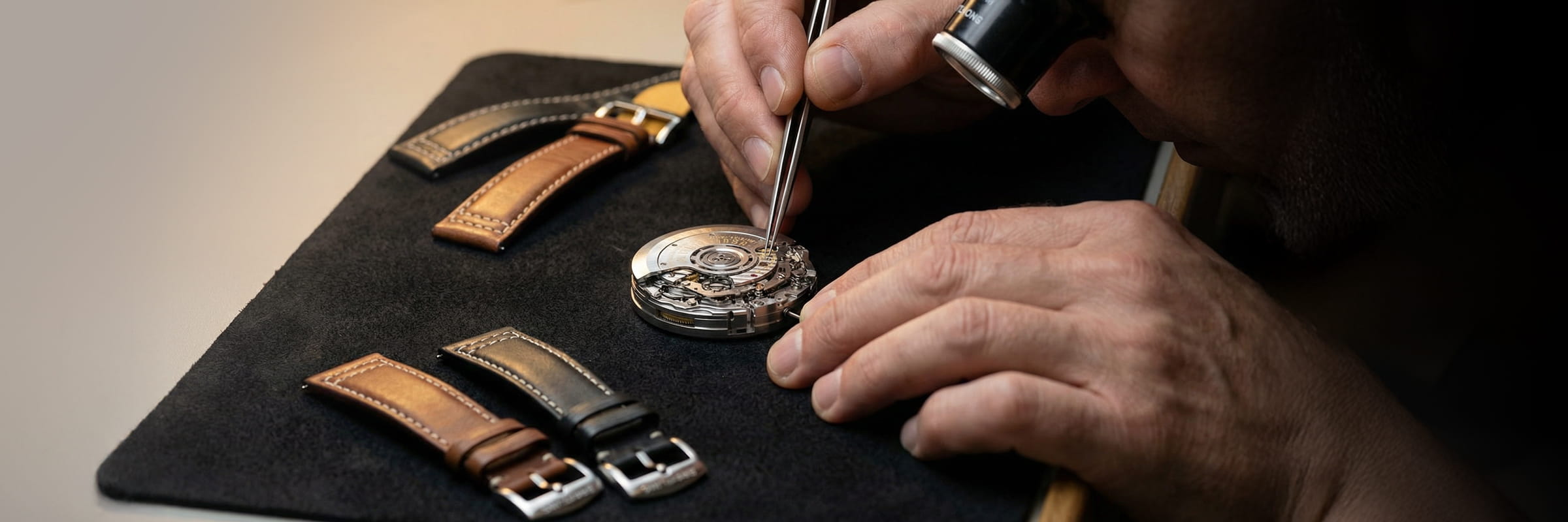 © Juwelier Wagner-Madler e.K. In einer Uhrenwerkstatt arbeitet ein Uhrmacher mit Lupe und Pinzette an einem Uhrwerk, während daneben mehrere Lederarmbänder auf einer schwarzen Matte ausgelegt sind.