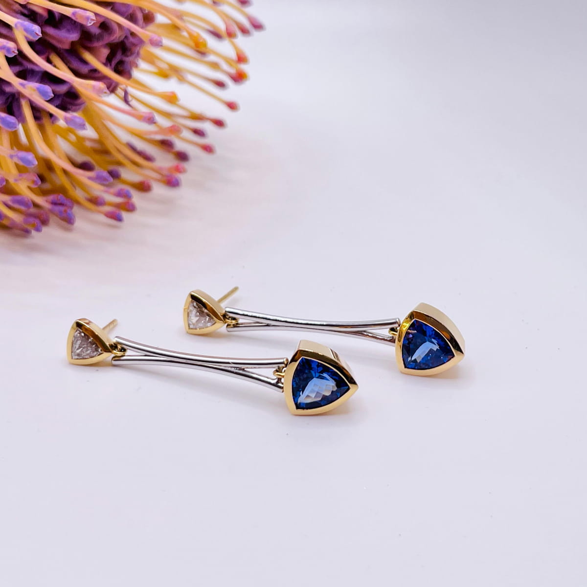 Ein Paar elegante Ohrringe mit blauen dreieckigen Edelsteinen in Goldfassungen, die durch glatte Silberstäbe mit kleineren klaren Steinen verbunden sind, die auf einer weißen Fläche neben einer stacheligen violetten und gelben Blume stehen.