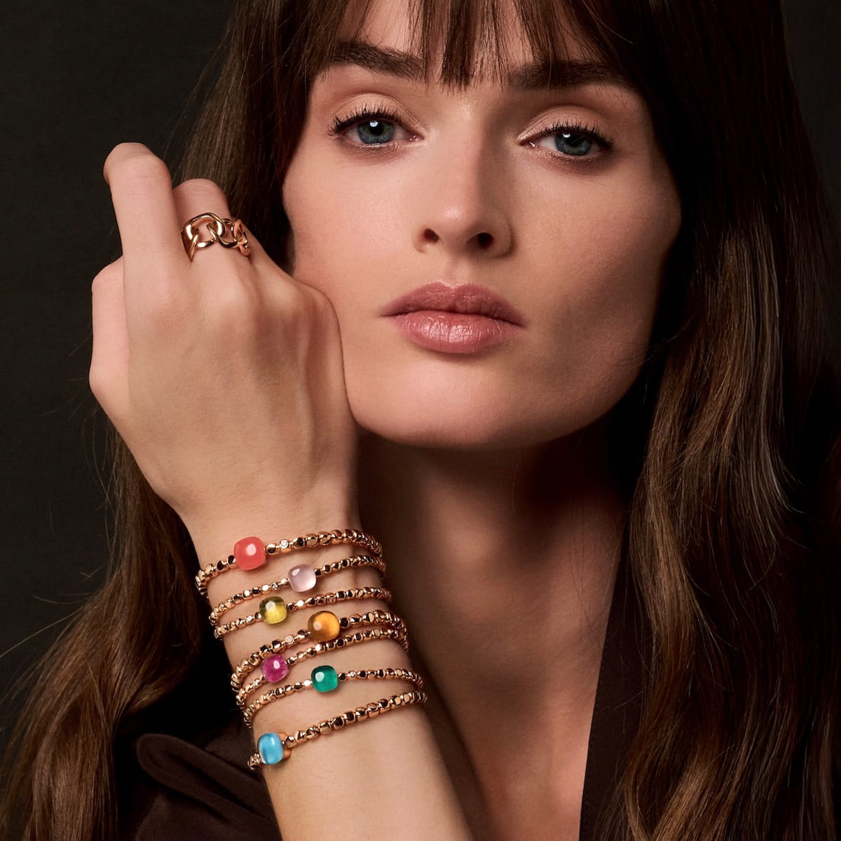 Eine Frau mit langem braunem Haar und Pony posiert mit der Hand in der Nähe ihres Gesichts und präsentiert goldene Bigli-Armbänder mit bunten Edelsteinen und einen goldenen Ring. Sie trägt ein dunkelbraunes Oberteil vor einem dunklen Hintergrund.