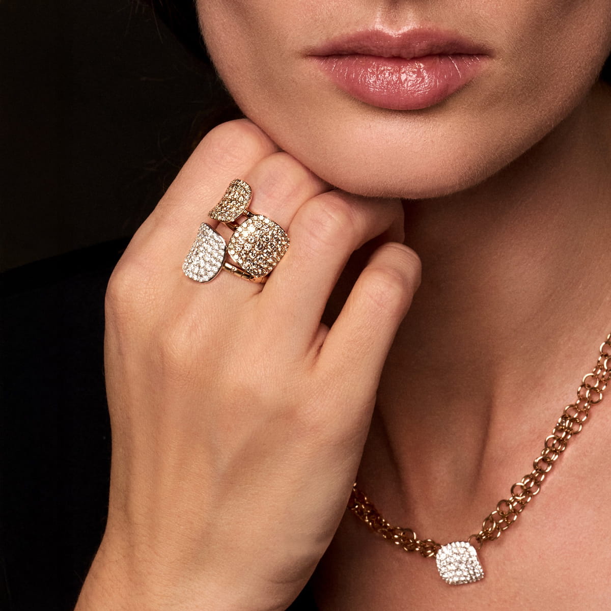 Die Hand einer Frau mit zwei Bigli-Gold- und Diamantringen ruht auf ihrem Kinn. Sie trägt eine passende goldene Halskette mit einem diamantbesetzten Anhänger vor dunklem Hintergrund.