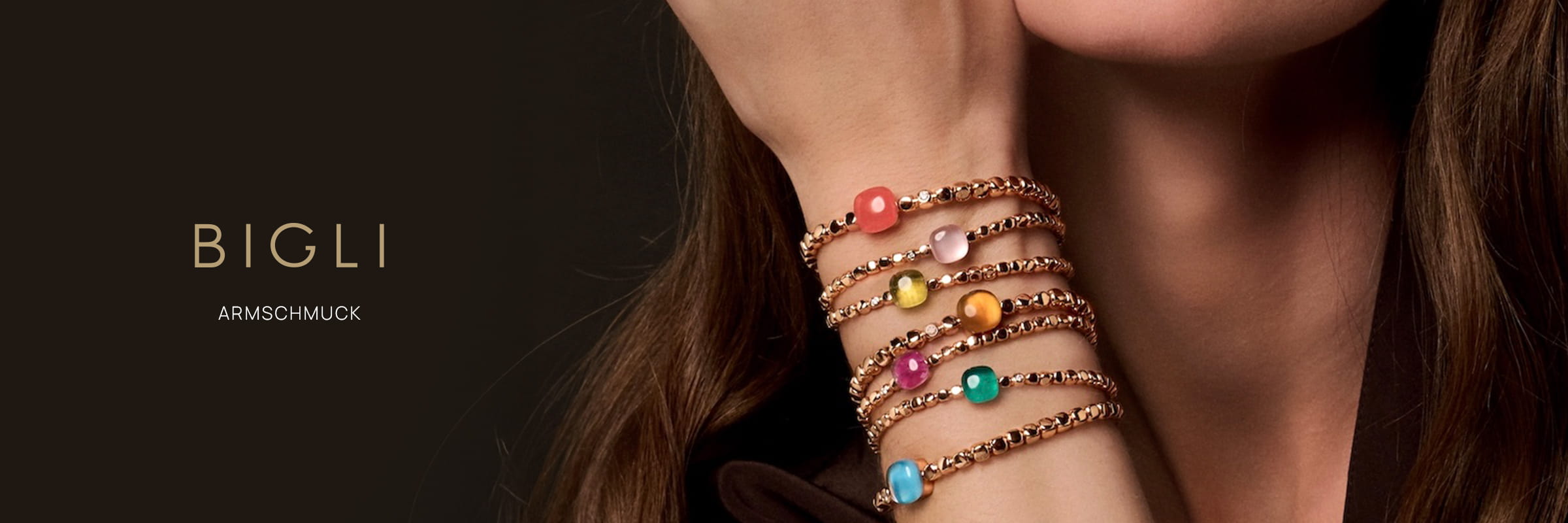 Das Handgelenk einer Frau ist mit mehreren goldenen Bigli-Armschmuck-Armbändern geschmückt, die jeweils mit einem bunten Edelstein besetzt sind. Der dunkle Hintergrund hebt den Text "BIGLI" und "ARMSCHMUCK" auf der linken Seite hervor.