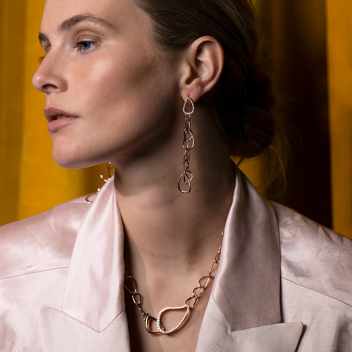 Eine Frau in einem blassrosa Satinblazer posiert vor einem goldenen Vorhang und trägt einen exquisiten Bigli-Halsschmuck - einen langen goldenen Kettenohrring und eine passende Halskette mit großen, sich überlappenden Schlaufen. Sie blickt mit ruhiger Miene zur Seite.