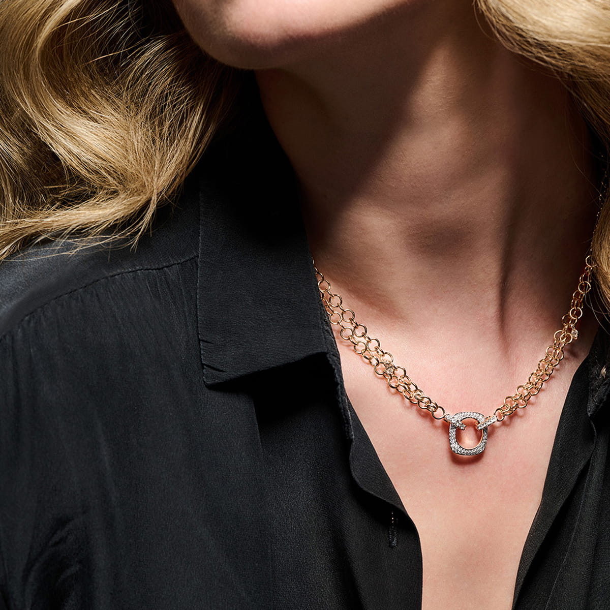 Eine Frau in schwarzer Bluse trägt eine goldene Halskette von Bigli Halsschmuck mit einem funkelnden, quadratischen Anhänger. Der Fokus liegt auf ihrem Hals und ihrem Schmuck, wobei ihr Gesicht teilweise aus dem Rahmen fällt und ihr gewelltes blondes Haar sichtbar ist.