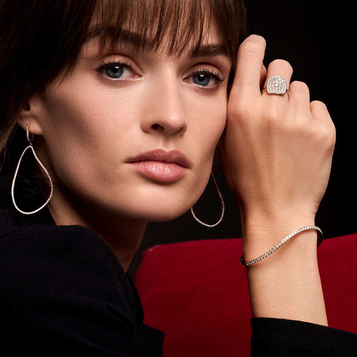 Eine Frau mit glattem braunem Haar trägt atemberaubende Bigli Ohrschmuck-Tropfenohrringe, einen Diamantring und ein Armband. Sie blickt mit auf die Hand gestütztem Kinn in die Kamera und sitzt vor einem rot-schwarzen Hintergrund.