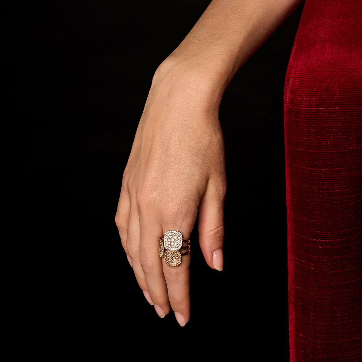 Eine Frauenhand mit sauber manikürten Nägeln ruht auf einer reichhaltigen roten Samtoberfläche und trägt drei gestapelte Bigli Ringe, die mit funkelnden Diamanten und Edelsteinen verziert sind. Der Hintergrund ist schwarz.