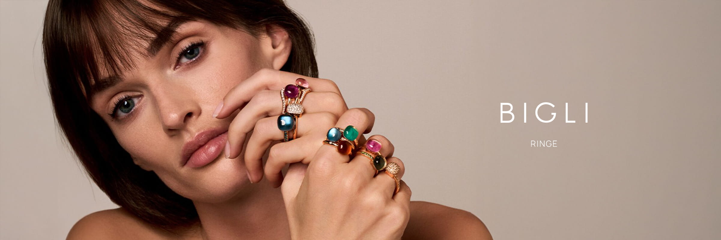 Eine Frau mit kurzen braunen Haaren posiert mit den Händen vor dem Gesicht und präsentiert den leuchtenden Edelstein Bigli Ringe. Der Hintergrund ist neutral und der Text lautet "BIGLI RINGE".