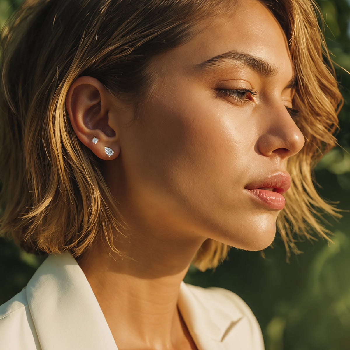 Eine Frau mit kurzem, gewelltem blondem Haar und leuchtender Haut trägt einen weißen Diavon-Blazer und kleine silberne Ohrstecker. Sie steht im Freien im Sonnenlicht vor einem verschwommenen grünen Hintergrund.