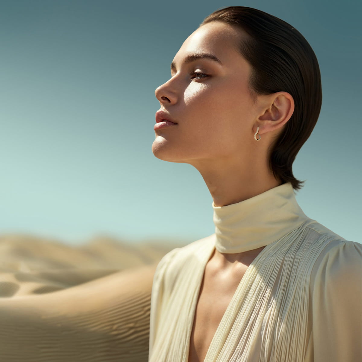 Eine Frau mit zurückgekämmtem Haar und einem cremefarbenen, hochgeschlossenen Diavon-Kleid steht in einer sonnenbeschienenen Wüste, die Augen geschlossen und den Kopf leicht nach oben geneigt, wobei das sanfte Sonnenlicht ihren heiteren Ausdruck hervorhebt.