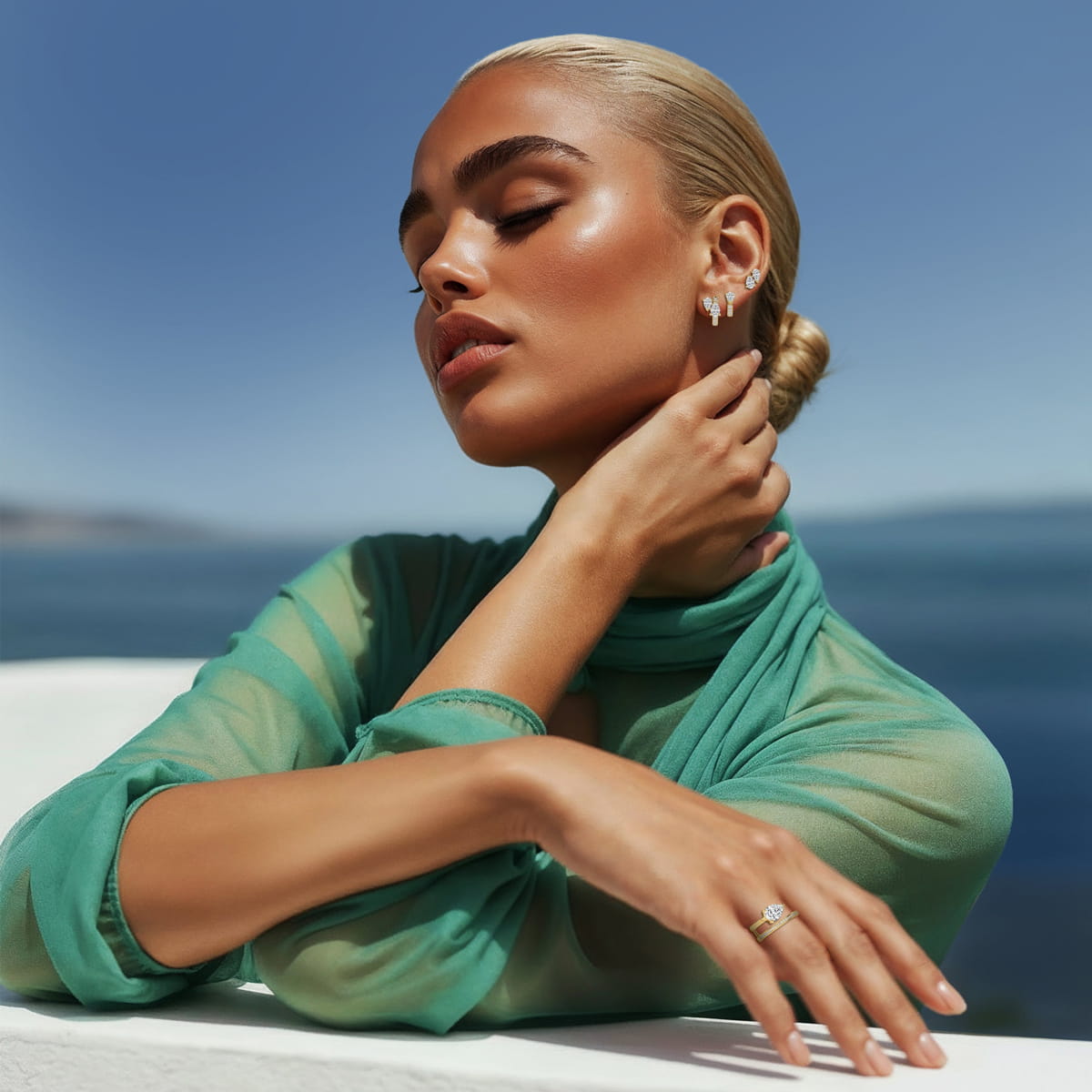 Eine Frau mit zurückgekämmtem blondem Haar posiert anmutig im Freien am Meer, trägt ein durchsichtiges grünes Oberteil und eleganten Schmuck, darunter Diavon Armschmuck, Ohrringe und einen Ring, vor einem klaren blauen Himmel und Wasserhintergrund.