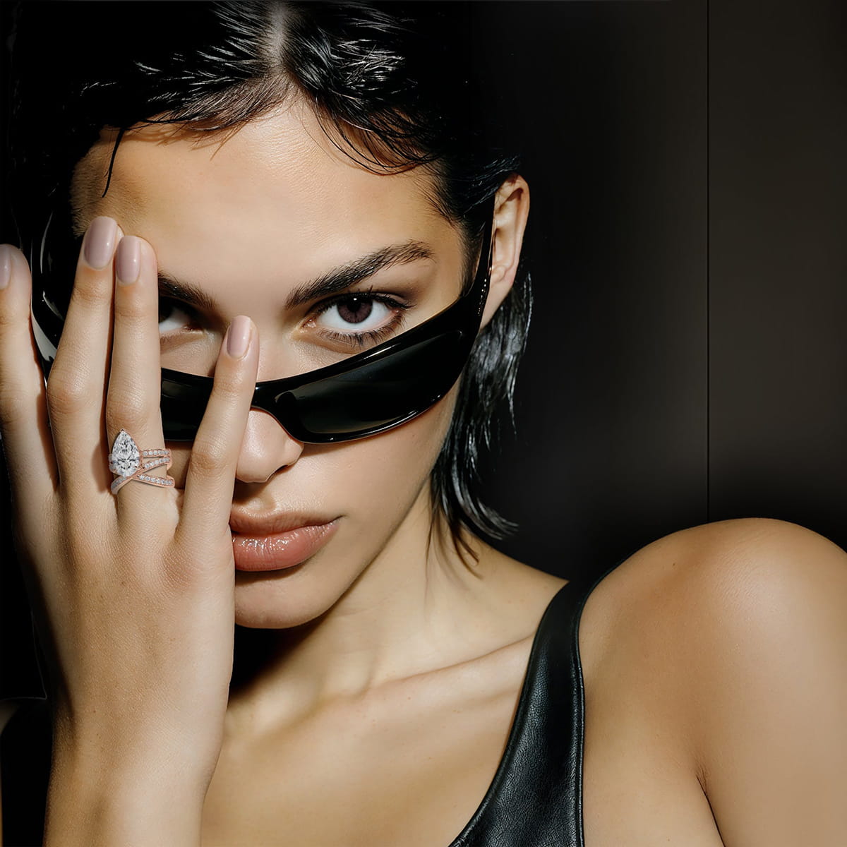 Eine Frau mit nassem, zurückgekämmtem Haar trägt eine schwarze Sonnenbrille und ein schwarzes Lederoberteil. Sie hält eine Hand vor ihr Gesicht und präsentiert einen großen, funkelnden Diamantring von Diavon Ringe. Der Hintergrund ist dunkel und unscharf.