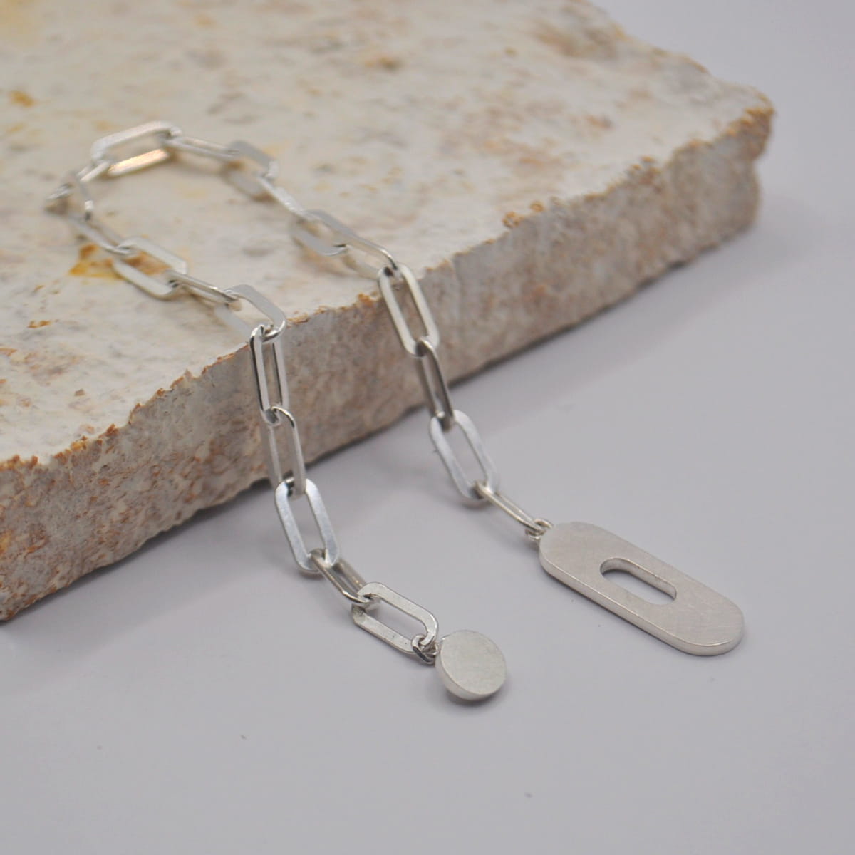 Silberne Halskette von DOPPELLUDWIG mit großen rechteckigen Gliedern, mit einem flachen ovalen Anhänger und einem flachen rechteckigen Anhänger, die auf einer rauen Steinplatte vor einem hellen Hintergrund angebracht sind.