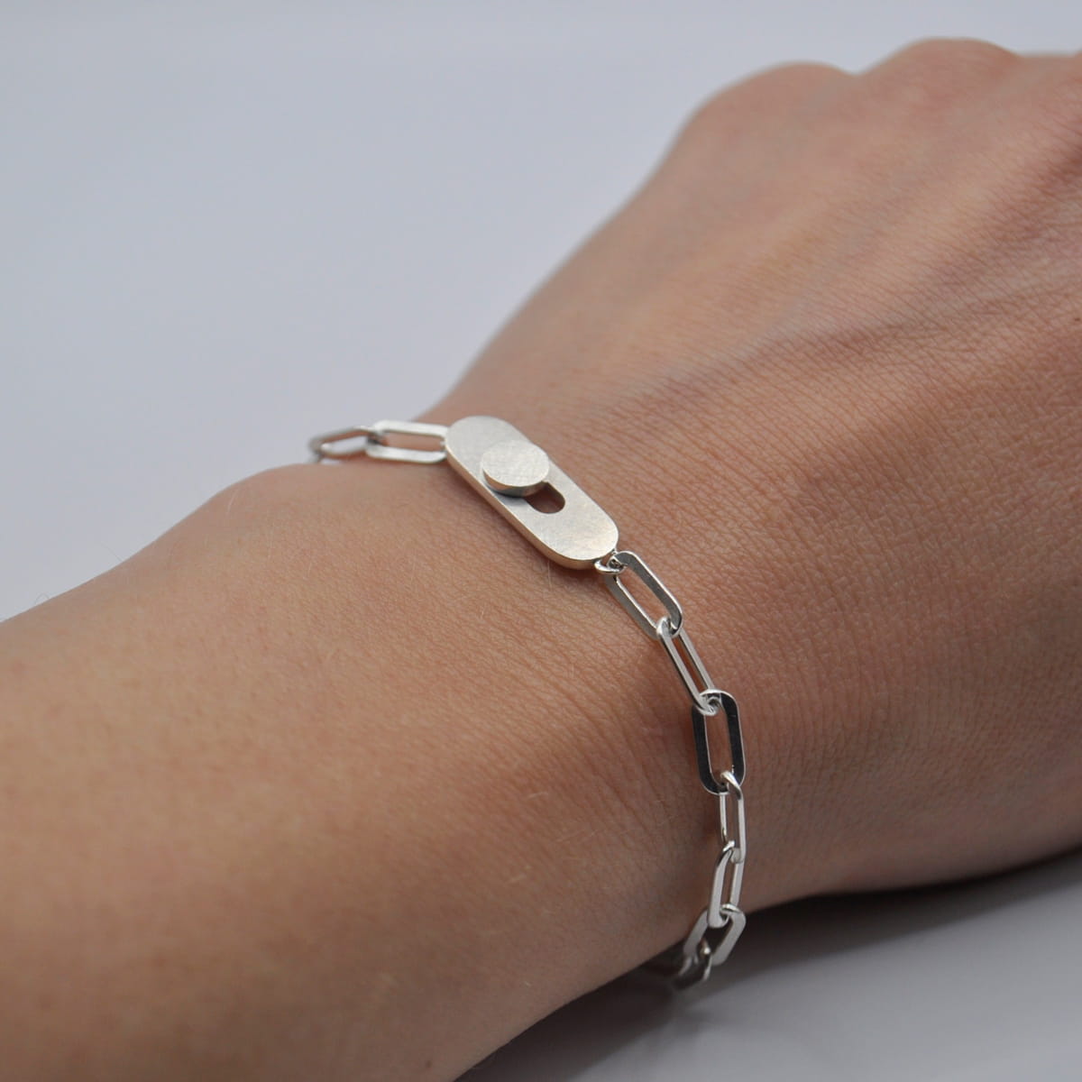 DOPPELLUDWIG © 2026 Nahaufnahme einer Hand, die ein silbernes Kettenarmband von DOPPELLUDWIG Armschmuck mit einem ovalen, strukturierten Mittelstück und länglichen Kettengliedern vor einem schlichten weißen Hintergrund trägt.