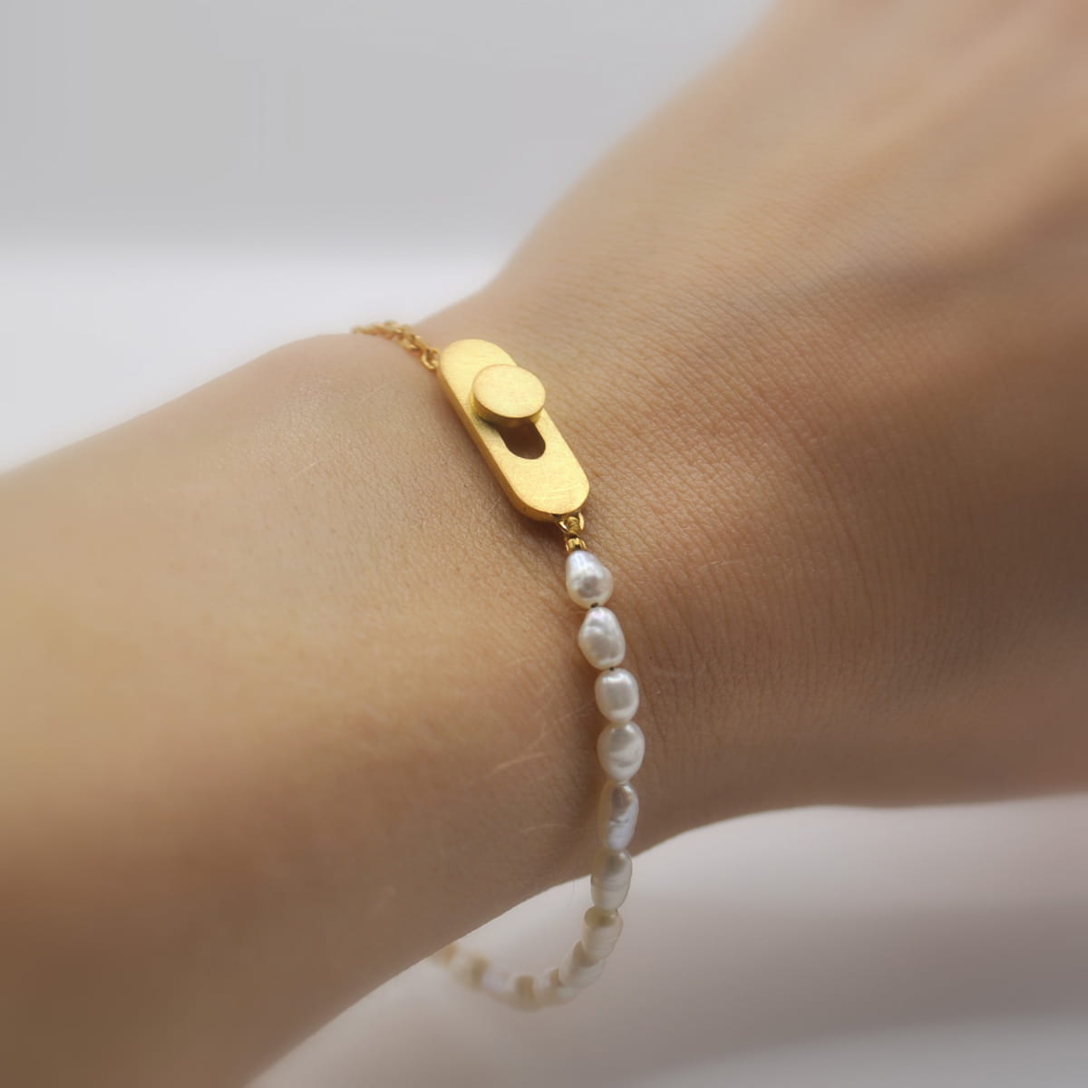 DOPPELLUDWIG © 2026 Nahaufnahme eines Handgelenks, das ein zartes Armschmuck-Armband von DOPPELLUDWIG trägt, das zur Hälfte aus einer Goldkette und zur Hälfte aus kleinen weißen Perlen besteht und einen einzigartigen Goldverschluss mit kreisförmigem und ovalem Muster aufweist.