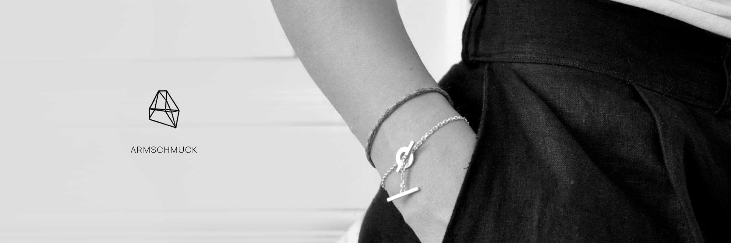 DOPPELLUDWIG © 2026 Eine Person mit DOPPELLUDWIG Armschmuck - ein Armband und eine Kette mit T-Verschluss -, gekleidet in einen schwarzen Rock und ein weißes Oberteil, die Hand in der Tasche. Das Wort "ARMSCHMUCK" und ein geometrisches Logo erscheinen auf der linken Seite.