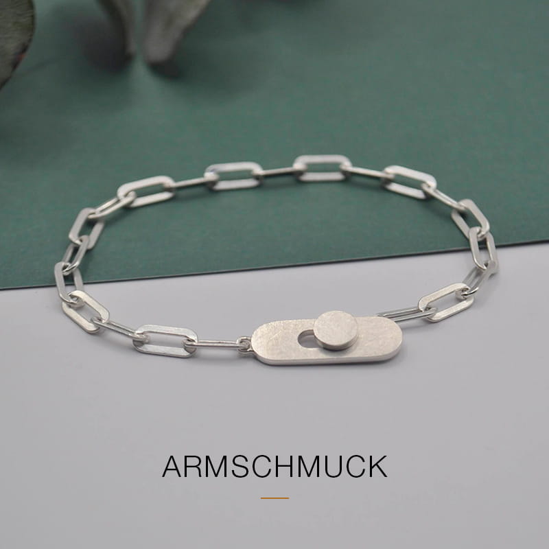 Ein minimalistisches silbernes Kettenarmband von DOPPELLUDWIG mit rechteckigen Gliedern und einem modernen Verschluss auf grünem und grauem Hintergrund. Der Schriftzug "ARMSCHMUCK" erscheint unterhalb des Armbands.