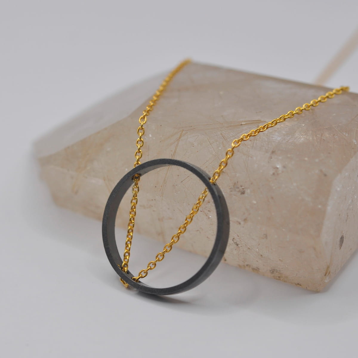 Eine goldene Halskette von DOPPELLUDWIG Halsschmuck mit einem schlichten, großen, schwarzen, runden Anhänger, der über einem durchscheinenden, rechteckigen Kristall auf weißem Grund drapiert ist.