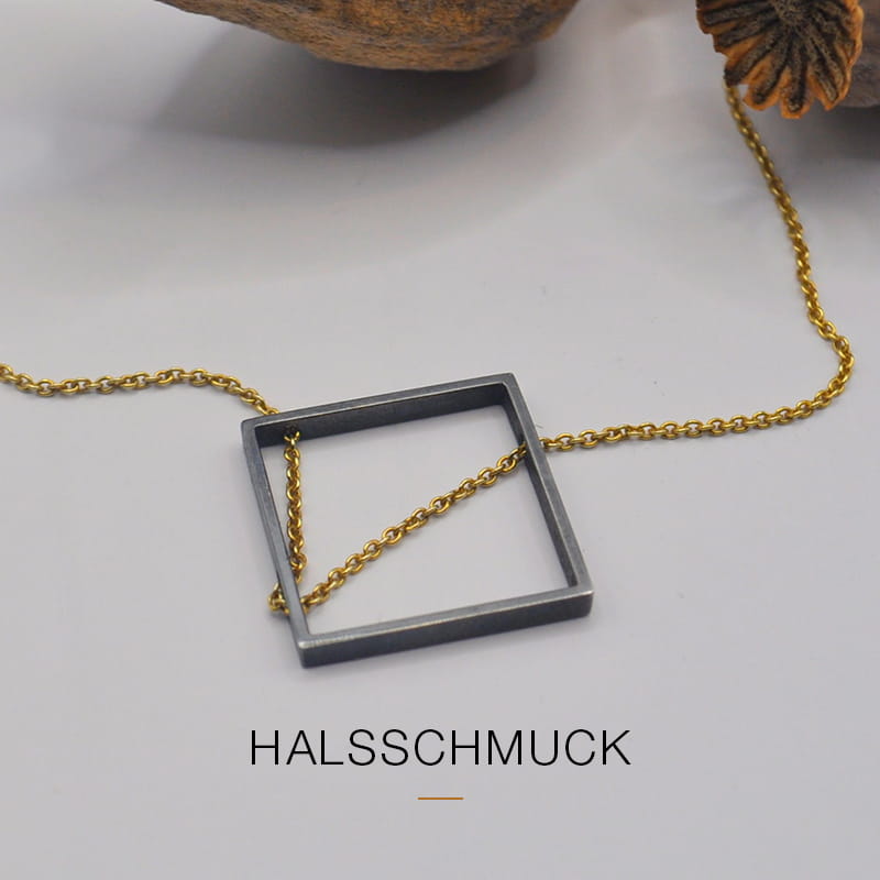 Eine goldene Halskette mit einem quadratischen, dunkelmetallischen DOPPELLUDWIG-Anhänger ist auf einer hellen Fläche ausgestellt. Unter der Halskette steht das Wort "HALSSCHMUCK".