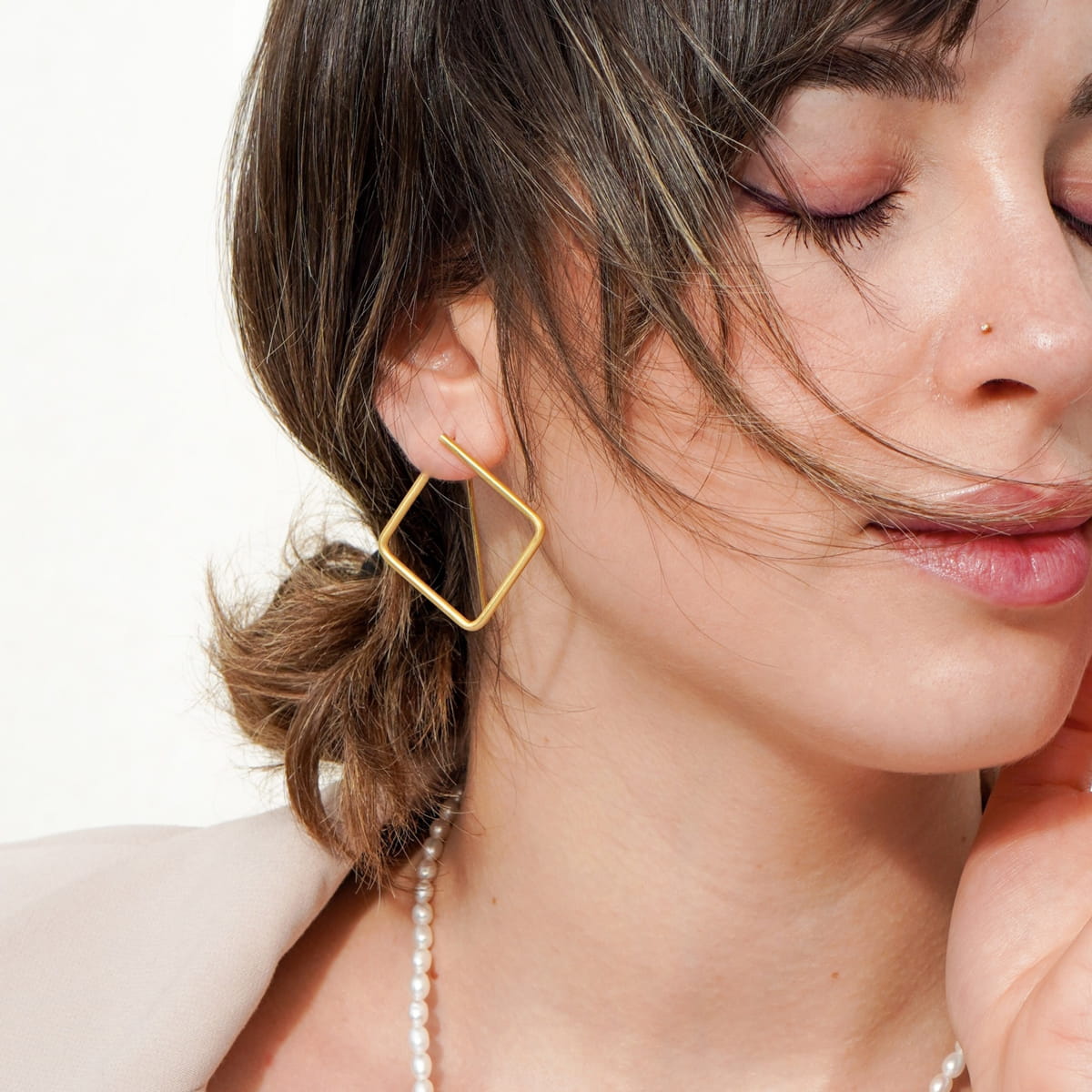 DOPPELLUDWIG © 2026 Eine Frau mit geschlossenen Augen und friedlichem Gesichtsausdruck trägt DOPPELLUDWIG Ohrschmuck - einen geometrischen Ohrring aus Gold - zusammen mit einer Perlenkette und einem Nasenstecker. Hellbraunes Haar umrahmt sanft ihr Gesicht.