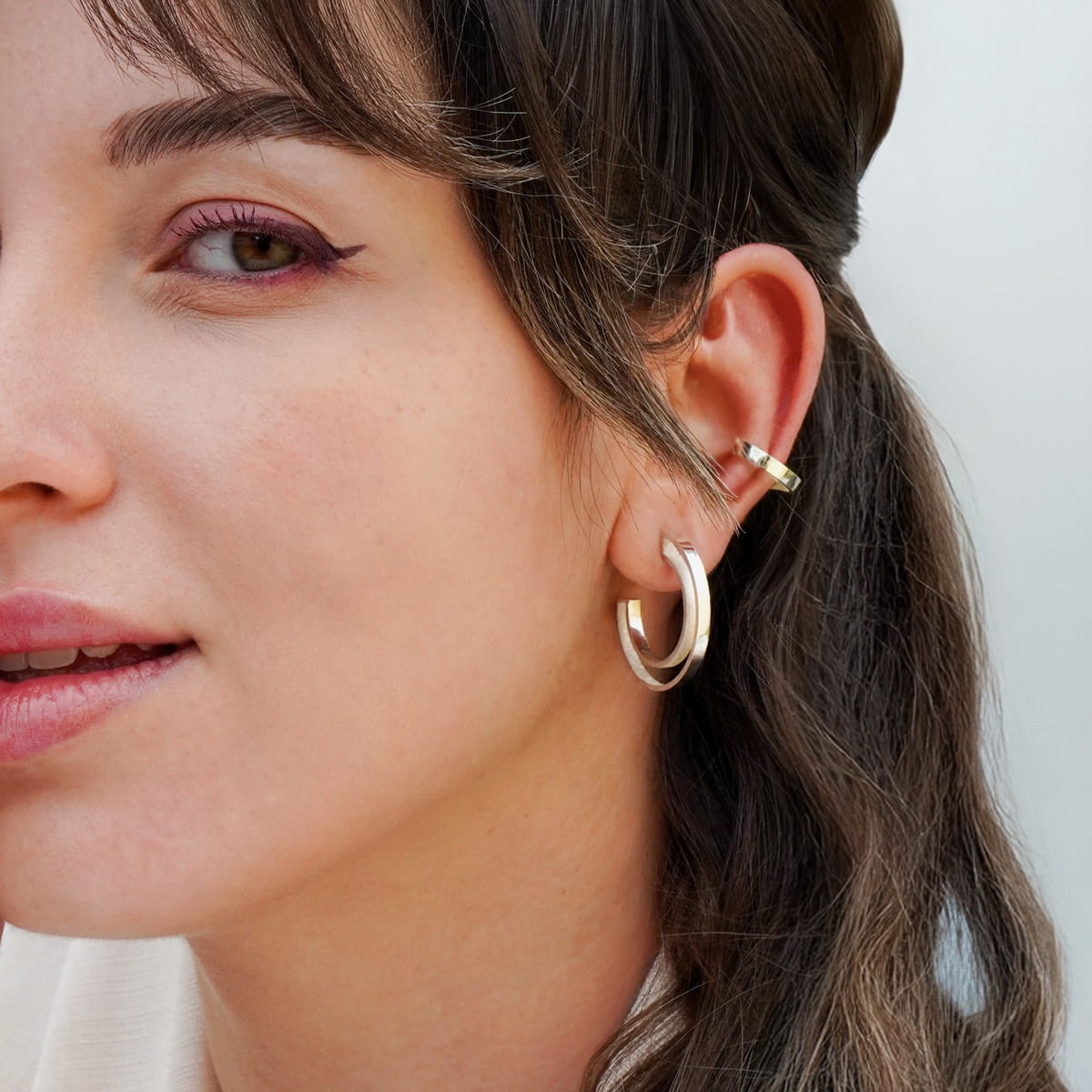 DOPPELLUDWIG © 2026 Nahaufnahme einer Frau mit heller Haut und braunem Haar, die DOPPELLUDWIG-Ohrschmuck in Gold und eine goldene Ohrmanschette trägt. Sie hat ein dezentes Make-up mit rosafarbenem Eyeliner und blickt leicht nach links und lächelt sanft.