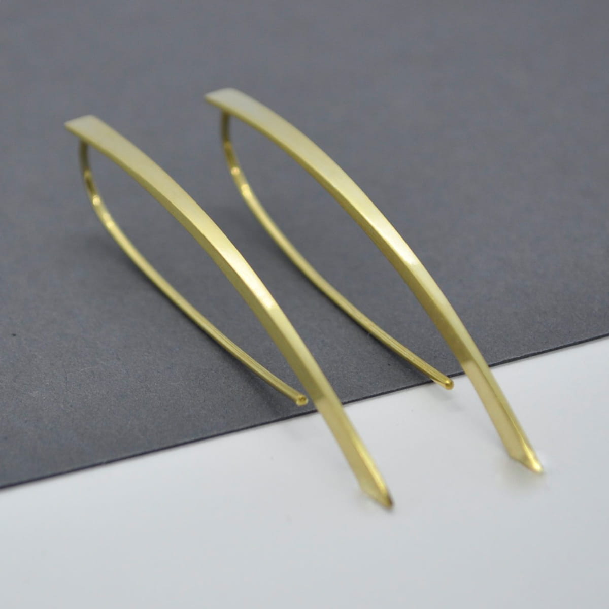 Ein Paar schlichte, minimalistische DOPPELLUDWIG-Ohrringe aus Gold mit geschwungenem, länglichem Design auf einem zweifarbigen Hintergrund in Grau und Weiß.